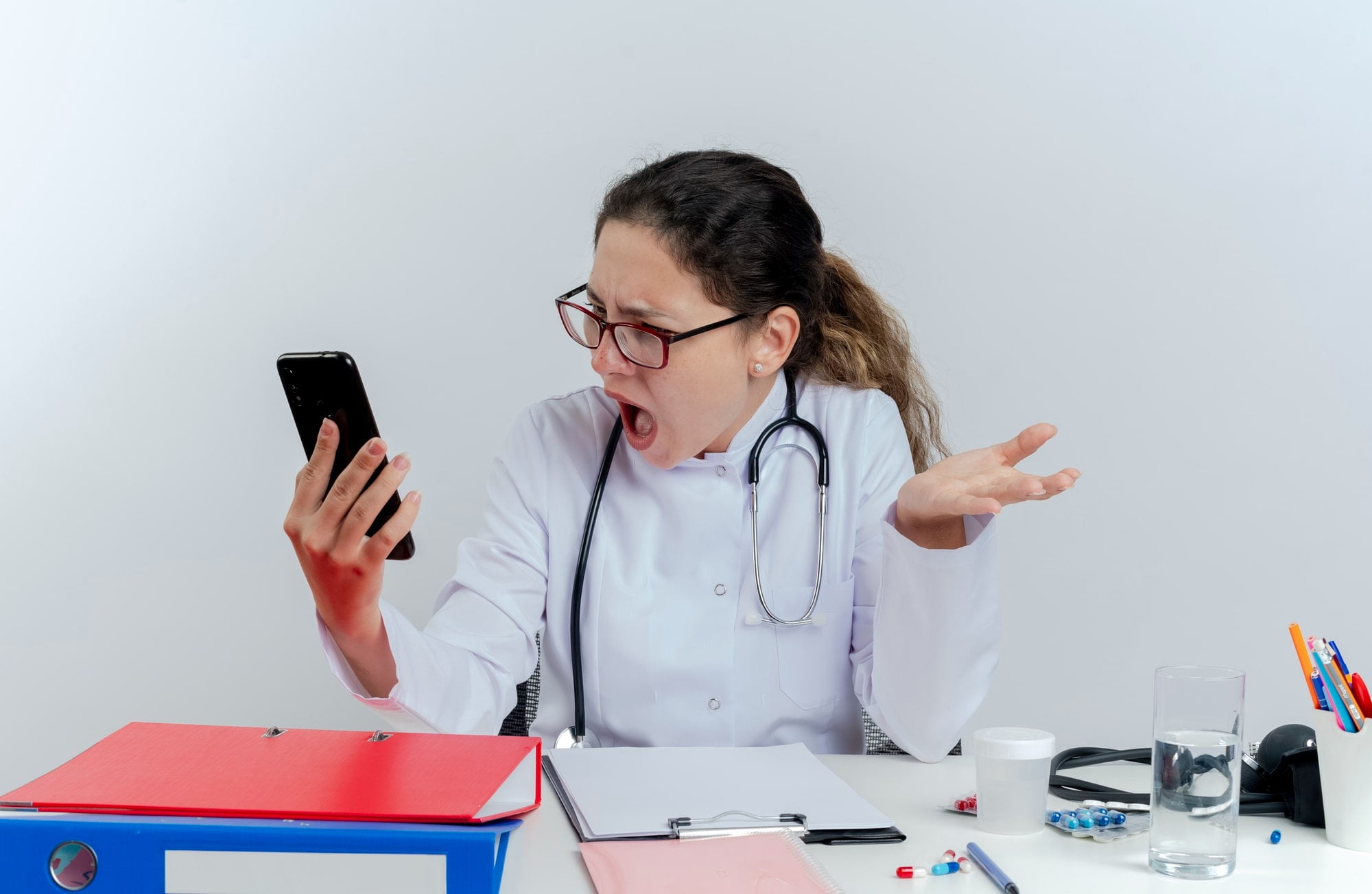 angry young female doctor wearing medical robe stethoscope glasses sitting desk with medical tools holding looking mobile phone showing empty hand isolated min